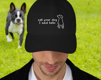 May include: Black baseball cap with the text "tell your dog I said hello" and a dog illustration in white. A Boston Terrier dog is in the background on a green lawn.