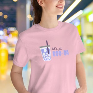 May include: A light pink t-shirt featuring a cartoon graphic of a bubble tea drink with ghost-shaped boba. The text "let's get BOO-BA" is printed in purple. The shirt is a crew neck style.