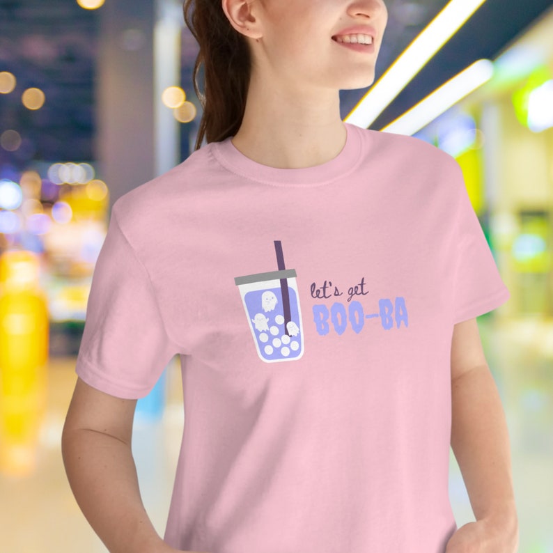 May include: A light pink t-shirt featuring a cartoon graphic of a bubble tea drink with ghost-shaped boba. The text "let's get BOO-BA" is printed in purple. The shirt is a crew neck style.
