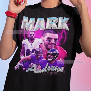 Op de afbeelding: Zwart T-shirt met een afbeelding van drie footballspelers in paarse en zwarte uniformen. De tekst "MARK ANDREWS" is gedrukt in een blauw en wit lettertype met een bliksemontwerp.