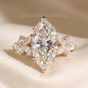 May include: A gold ring featuring a large marquise-cut center stone held by prongs, flanked by two clusters of smaller marquise and round-cut stones on each side. The text GOLDMOONJEWELS is visible above the ring, which has a polished gold band.