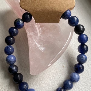May include: Sodalite beaded bracelet with varying shades of blue and black beads. The bracelet is displayed on a pink, heart-shaped object. A brown tag with the word "Sodalite" is attached.