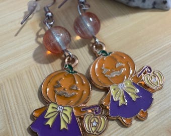 May include: A pair of Halloween-themed earrings. Each earring features a smiling orange pumpkin character wearing a purple dress and holding a small lantern. The earrings have orange and clear bead accents and silver-colored ear wires.