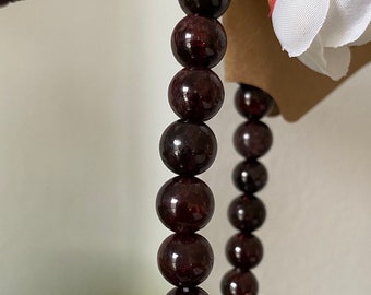 May include: A dark red gemstone bracelet made of round beads. The bracelet is strung on a clear elastic cord.