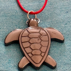 May include: A black hematite turtle pendant on a red cord necklace. The turtle is carved with a detailed shell pattern.