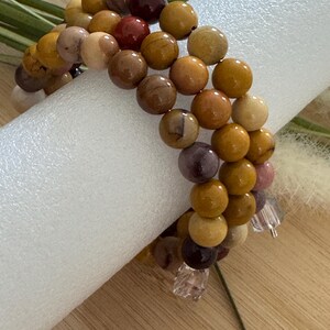 Könnte beinhalten: Ein mehrsträngiges Armband mit runden, polierten Perlen in Braun-, Beige- und Burgundertönen. Die Perlen sind aufgereiht, mit klaren, facettierten Kristallakzenten. Das Armband wird auf einer weißen zylindrischen Form präsentiert.