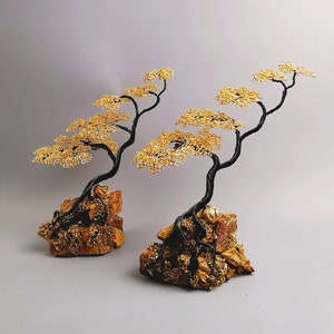 May include: Two wire bonsai trees with black trunks and gold wire foliage. The trees are mounted on textured, golden-brown rock bases. The bonsai trees are a decorative item.