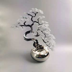 May include: A wire sculpture of a bonsai tree featuring a twisted black and silver trunk with delicate silver wire foliage. The tree is anchored in a round, polished chrome pot filled with smooth white stones and small pebbles.
