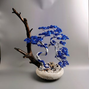 May include: A blue and silver wire bonsai tree with a brown wooden base, sitting in a white ceramic pot with white stones and seashells.