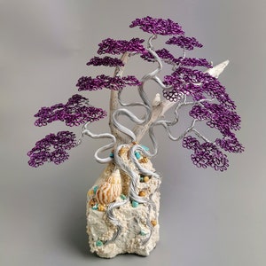 May include: A purple wire tree sculpture with a white base and seashells. The tree is made of wire and has a natural, organic look. The base is made of a white material and has a rough texture. The seashells are a natural addition to the sculpture and add a touch of whimsy.
