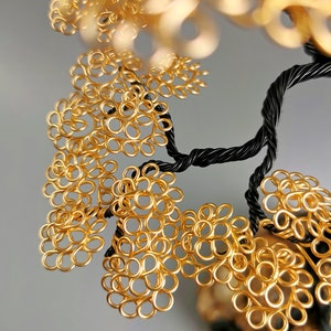 May include: A handcrafted wire tree sculpture with a black twisted trunk and branches. The tree's leaves are made of gold-colored wire, creating a textured, ornamental design. The sculpture is set against a soft gray background.