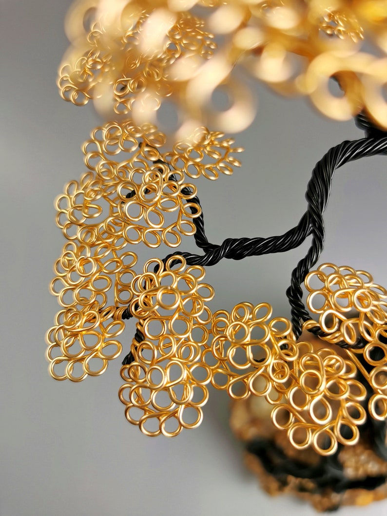 May include: A handcrafted wire tree sculpture with a black twisted trunk and branches. The tree's leaves are made of gold-colored wire, creating a textured, ornamental design. The sculpture is set against a soft gray background.