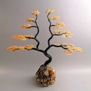 May include: A handcrafted wire tree sculpture with a black twisted trunk and branches, adorned with gold-colored wire leaves. The tree is rooted in a textured, brown stone base, creating a miniature bonsai aesthetic.