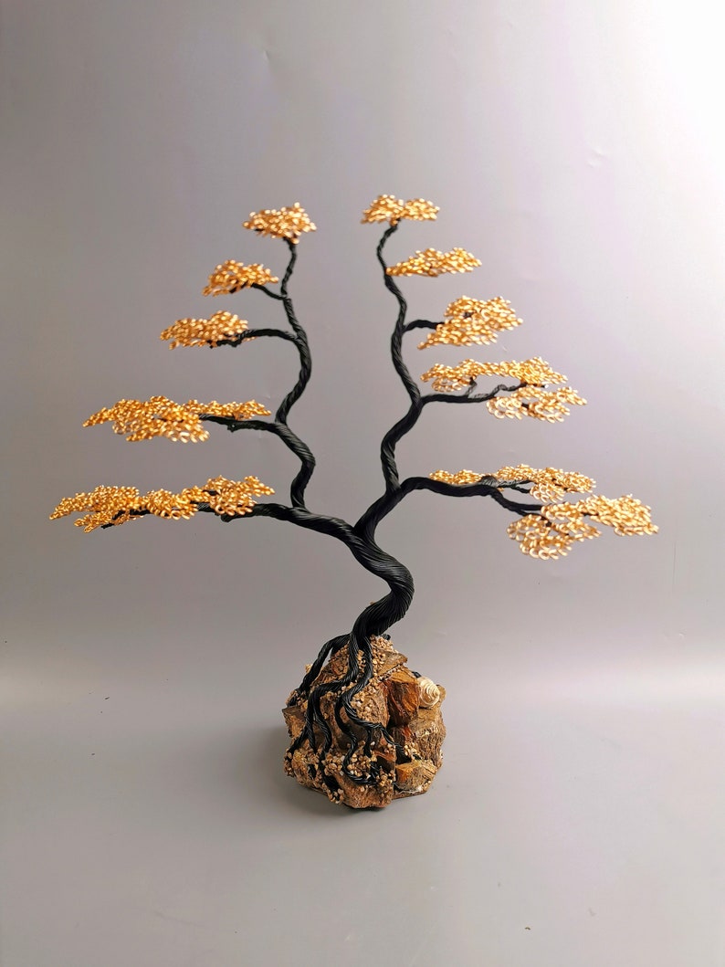 May include: A handcrafted wire tree sculpture with a black twisted trunk and branches, adorned with gold-colored wire leaves. The tree is rooted in a textured, brown stone base, creating a miniature bonsai aesthetic.