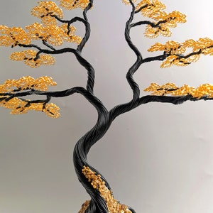 May include: A handcrafted wire tree sculpture with a black twisted trunk and branches. The tree's leaves are a vibrant gold color. The base of the tree is a rock formation with small pebbles.