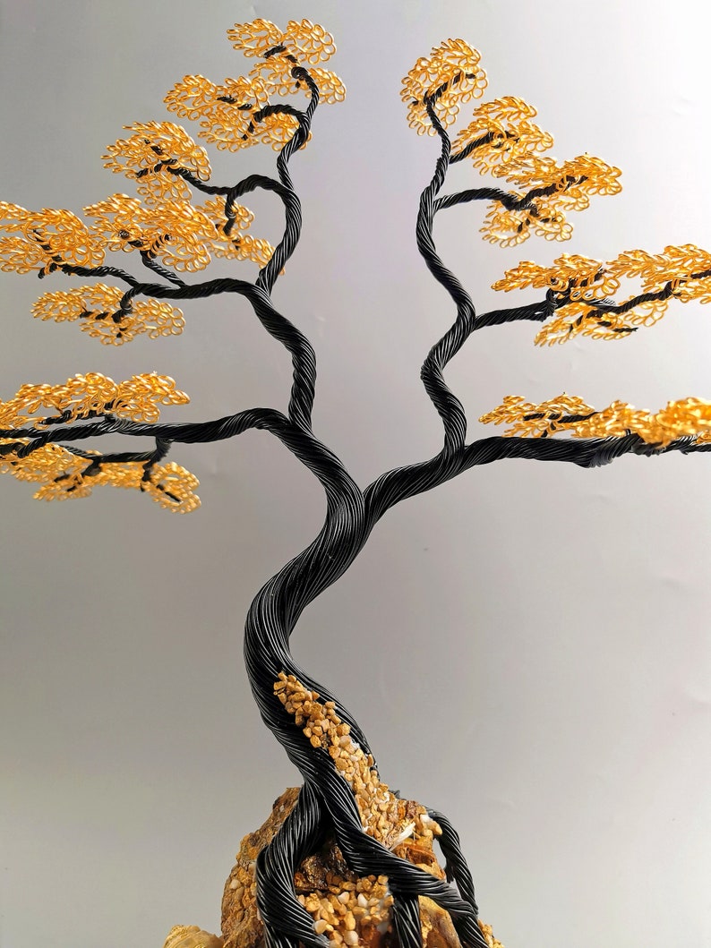 May include: A handcrafted wire tree sculpture with a black twisted trunk and branches. The tree's leaves are a vibrant gold color. The base of the tree is a rock formation with small pebbles.