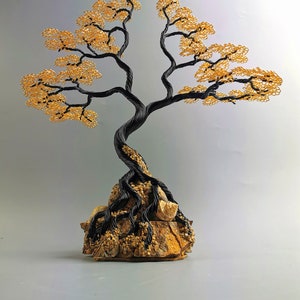 May include: A handcrafted wire tree sculpture with a black trunk and branches, and golden-yellow leaves. The tree is rooted in a textured, tan-colored stone base, creating a miniature bonsai-like aesthetic.