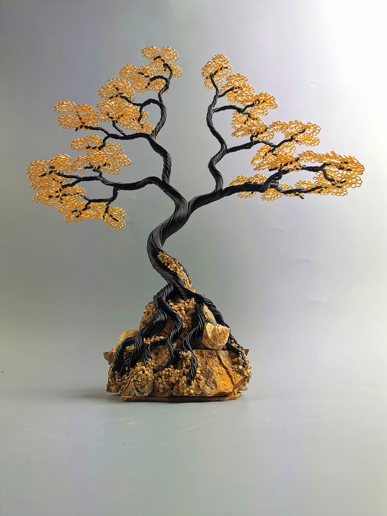 May include: A handcrafted wire tree sculpture with a black trunk and branches, and golden-yellow leaves. The tree is rooted in a textured, tan-colored stone base, creating a miniature bonsai-like aesthetic.