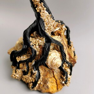 May include: A handcrafted sculpture featuring a black wire tree with intricate roots, adorned with small, golden-colored stones and a seashell. The tree is mounted on a natural, textured rock base, creating a unique decorative piece.