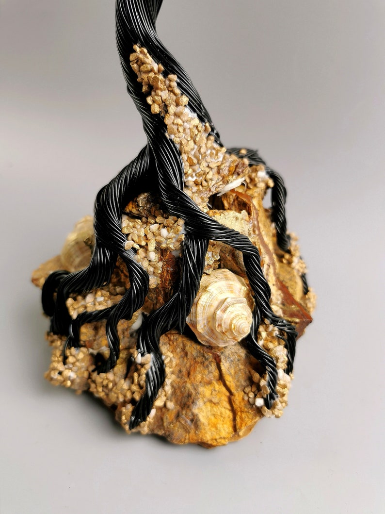 May include: A handcrafted sculpture featuring a black wire tree with intricate roots, adorned with small, golden-colored stones and a seashell. The tree is mounted on a natural, textured rock base, creating a unique decorative piece.