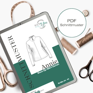 Könnte beinhalten: Ein digitales Tablet zeigt ein Schnittmuster für ein Reißverschluss-Shirt namens "Annie". Das Schnittmuster ist als PDF-Download erhältlich und ist für die Größen 34-44. Das Tablet liegt auf einer weißen Oberfläche mit Nähzubehör, darunter eine Schere, ein Maßband und Garnrollen.