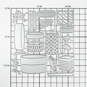 May include: A set of 13 metal die-cut shapes for crafting. The shapes include a variety of boxes, jars, bows, and other decorative elements. The shapes are all silver in color and are arranged on a gridded background.