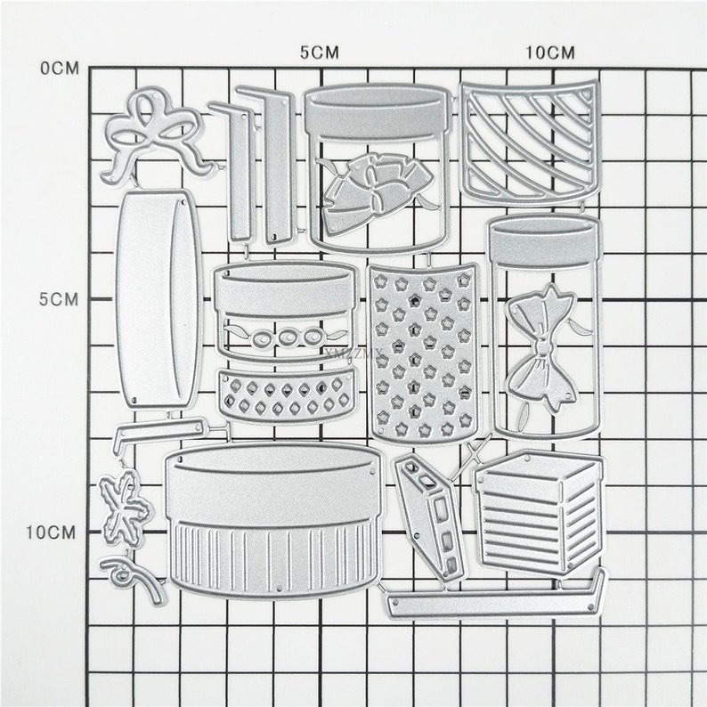 May include: A set of 13 metal die-cut shapes for crafting. The shapes include a variety of boxes, jars, bows, and other decorative elements. The shapes are all silver in color and are arranged on a gridded background.