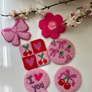 Könnte beinhalten: Sechs rosa und rote Filz-Handarbeiten mit verschiedenen Designs. Die Designs umfassen eine Schleife, eine Blume mit einem Smiley, ein kariertes Muster mit Herzen und Kirschen, eine Sprechblase mit dem Text "Ich liebe dich" und zwei Kreise mit Kirschen.