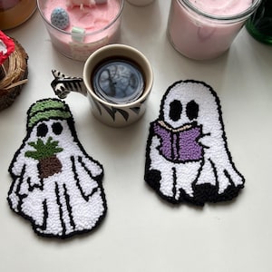 Op de afbeelding: Twee witte spookvormige onderzetters met zwarte contouren. Eén spook houdt een groene plant vast en de andere houdt een paars boek vast.