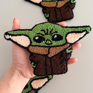 May include: Handmade green, brown, and beige embroidered patches featuring the character Grogu from The Mandalorian. The patches have large black eyes and are in a standing pose. The patches are made with a textured, looped stitch.