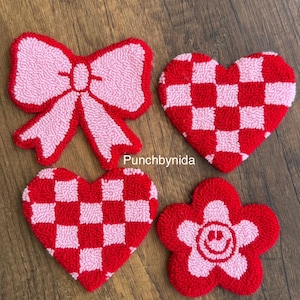 Op de afbeelding: Vijf handgemaakte vilt wanddecoraties in de vorm van een strik, een bloem en drie harten. De harten zijn geruit met rode en roze vierkanten. De bloem heeft een roze centrum met een smiley en rode bloemblaadjes. De strik is roze met rode accenten.