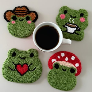 May include: Four green frog-shaped coasters with various designs, including a cowboy hat, a coffee cup, a heart, and a mushroom. A white mug filled with black coffee sits in the centre. The coasters are made of a textured material.