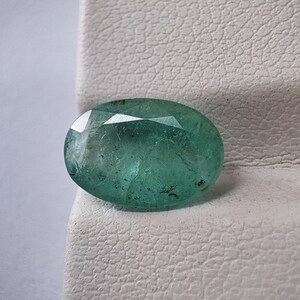 Pode incluir: Gema de esmeralda ovalada e facetada. A gema é de um verde profundo e translúcido com inclusões visíveis na pedra. O corte da esmeralda cria múltiplas superfícies refletoras, realçando o seu brilho.