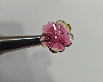 May include: A carved gemstone flower with pink and yellow hues is held by a metal tool. The flower has detailed petal carvings and a central hole. The gemstone is translucent, allowing light to pass through.