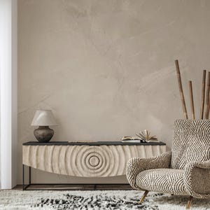 May include: A neutral-toned living room scene featuring a carved wooden cabinet with a black top, a patterned armchair, a lamp, and decorative bamboo stalks. The cabinet has a circular design on the doors. A rug with a black and white pattern is visible.