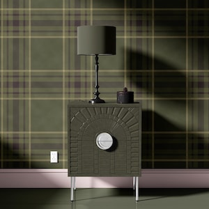 May include: A dark green nightstand with a decorative arched front and silver legs. A black lamp with a green lampshade sits on top, along with a small black box. The background features a green and brown plaid pattern.