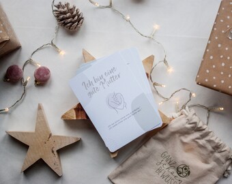 Könnte beinhalten: Ein Stapel weißer Karten mit dem deutschen Text "Ich bin eine gute Mutter" (Ich bin eine gute Mutter) liegt auf einem Holzstern. Das Bild enthält einen kleinen Kordelzugbeutel, verpackte Geschenke, Holzsterne und Lichterketten.