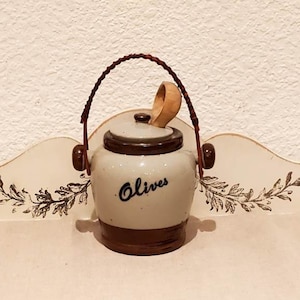 Puede incluir: Un tarro de cerámica para aceitunas con tapa marrón y cuchara de madera. El tarro tiene un asa marrón y la palabra "Olives" escrita en negro en la parte delantera.