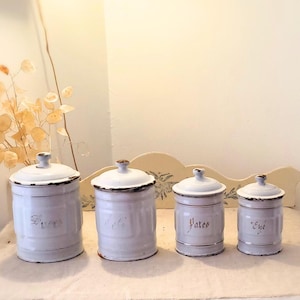 Peut inclure: Quatre pots en céramique blanche avec des inscriptions dorées sur une étagère blanche. Les pots sont étiquetés "Sucre", "The", "Pâtes" et "Café".