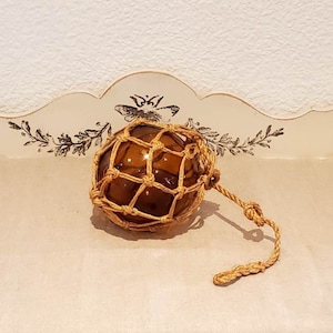 Può includere: Un galleggiante da pesca in vetro marrone con una rete di corda beige. Il galleggiante è su una superficie bianca.