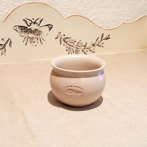 May include: A small, round, beige ceramic pot with a raised design of a building and the text "Céramique de Marseille".