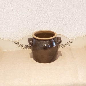 May include: A brown ceramic crock with two handles. The crock has a glazed finish and is sitting on a white surface.