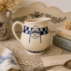 May include: A cream-colored ceramic pitcher featuring a blue and white checkered band around the top and base. The front displays a blue oval logo with the text Pâtes aux œufs frais Lustucru, fabriquees par les pâtes Lustucru, and a handle.