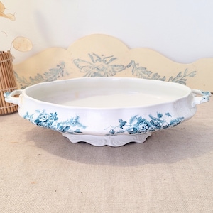 Pode incluir: Uma tigela de servir oval branca com duas alças e uma borda festonada. A tigela é decorada com um padrão floral azul.