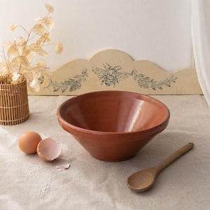 May include: A terracotta-colored mixing bowl with a wide rim sits on a linen surface. Two eggs, one cracked, and a wooden spoon are nearby. A decorative piece with a bee design is in the background.