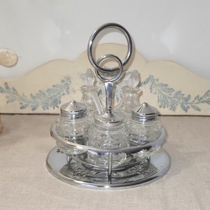 May include: A silver condiment set with five glass bottles and a circular base. The bottles have a textured glass design and are held in place by a silver frame. The set has a handle with two rings.