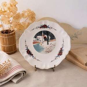 Op de afbeelding: Een witte decoratieve plaat met een geschulpte rand, met een winterse scène met een kerk en een rivier. De plaat is versierd met bloemaccenten en rust op een bronzen standaard. Een rustieke, geweven vaas en vintage boeken staan op de achtergrond.
