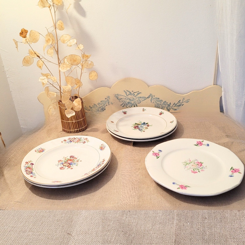 May include: Three white ceramic plates with floral designs. The plates are stacked on a light brown surface.