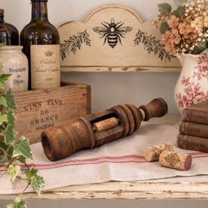 Pode incluir: Uma ferramenta de rolha para vinho em madeira castanho-escura com corpo cilíndrico e abertura central, acompanhada por duas pequenas rolhas com carimbos decorativos. A ferramenta tem um cabo de madeira torneada e uma estrutura robusta.