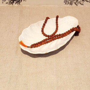 Puede incluir: Un plato de cer&aacute;mica blanco con forma de concha de mar con un collar de cuentas marrones en su interior.